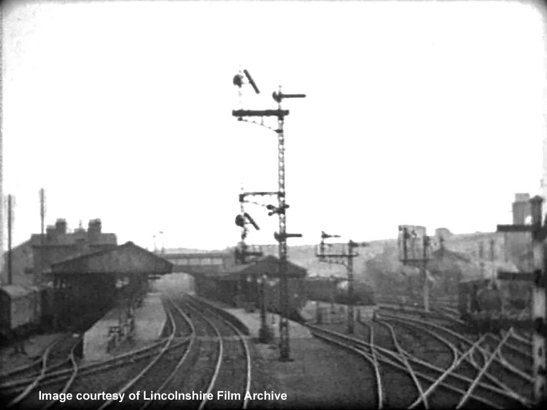 This is the Signalman’s view, looking south, of the station from Grantham North box in 1937. The most prominent signal post, at the centre of the view, controls the Down (northbound) Main line immediately to its right (as viewed from an approaching train). In this direction behind the photographer there is a choice of two routes: continuing on the Main line towards Newark, or a junction diverging to the left towards Nottingham (again, as viewed from an approaching train). A signal relating to the Nottingham line is therefore situated to the left of the Main line signal as viewed by an approaching driver. The Nottingham line signal is also at a slightly lower height because it refers to a less important route. This particular signal has duplicate ‘co-acting’ arms at high level because the station footbridge prevents drivers seeing the low level signals from a distance. So we can ‘read’, from the location of the inclined arms in the photograph, that the route is set for a train to pass northbound through the station and to continue along the Main line. To the right of the tall post, as viewed in the photograph, are two similar junction signals (without high level co-acting arms) which control northbound departures from the Bay platform and the Western platform. Both provide the option of Main line or Nottingham line routing. On the extreme right of the photograph is a signal post with three arms. This controls trains approaching the station from the Nottingham line. Each arm indicates a different route and they read ‘top to bottom’ = ‘left to right’. So far as we can tell (can anyone confirm or correct this?) if the top arm is lowered the route would be set into the Bay platform, the middle arm probably applies to the short engine spur off the Bay platform line, and the bottom arm most likely indicates the Western platform. This vertical arrangement of signal arms indicating different routes had been in use since the earliest days of railway signalling. Bracket-type junction signals gave a clearer indication and they had generally replaced the vertical arrangement, except in sidings. Photo taken from a cine film shot by photographer and film maker Walter Lee of Grantham. © Lincolnshire Film Archive